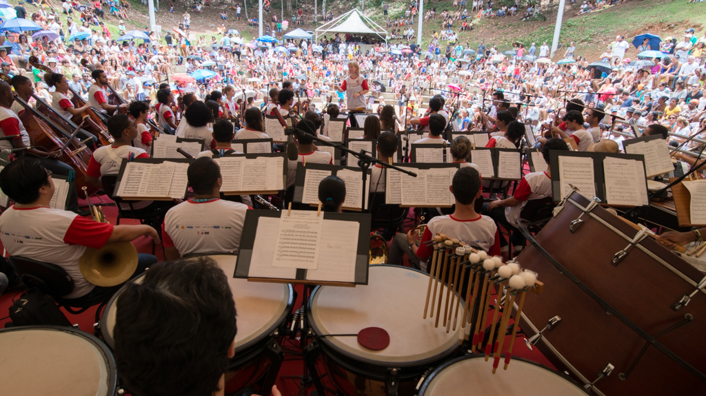 Imagem mostra apresentação da Orquestra baiana Neojiba, no Parque da Cidade, em Salvador. Em primeiro plano, os jovens músicos de costas, a maestrina de frente para eles regendo e, ao fundo, o público assistindo atento