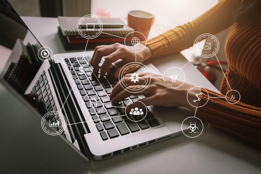 imagem mostra mãos femininas digitando no teclado de um notebook em cima de uma mesa, onde também se vê um caderno pequeno e uma caneca desfocados na imagem