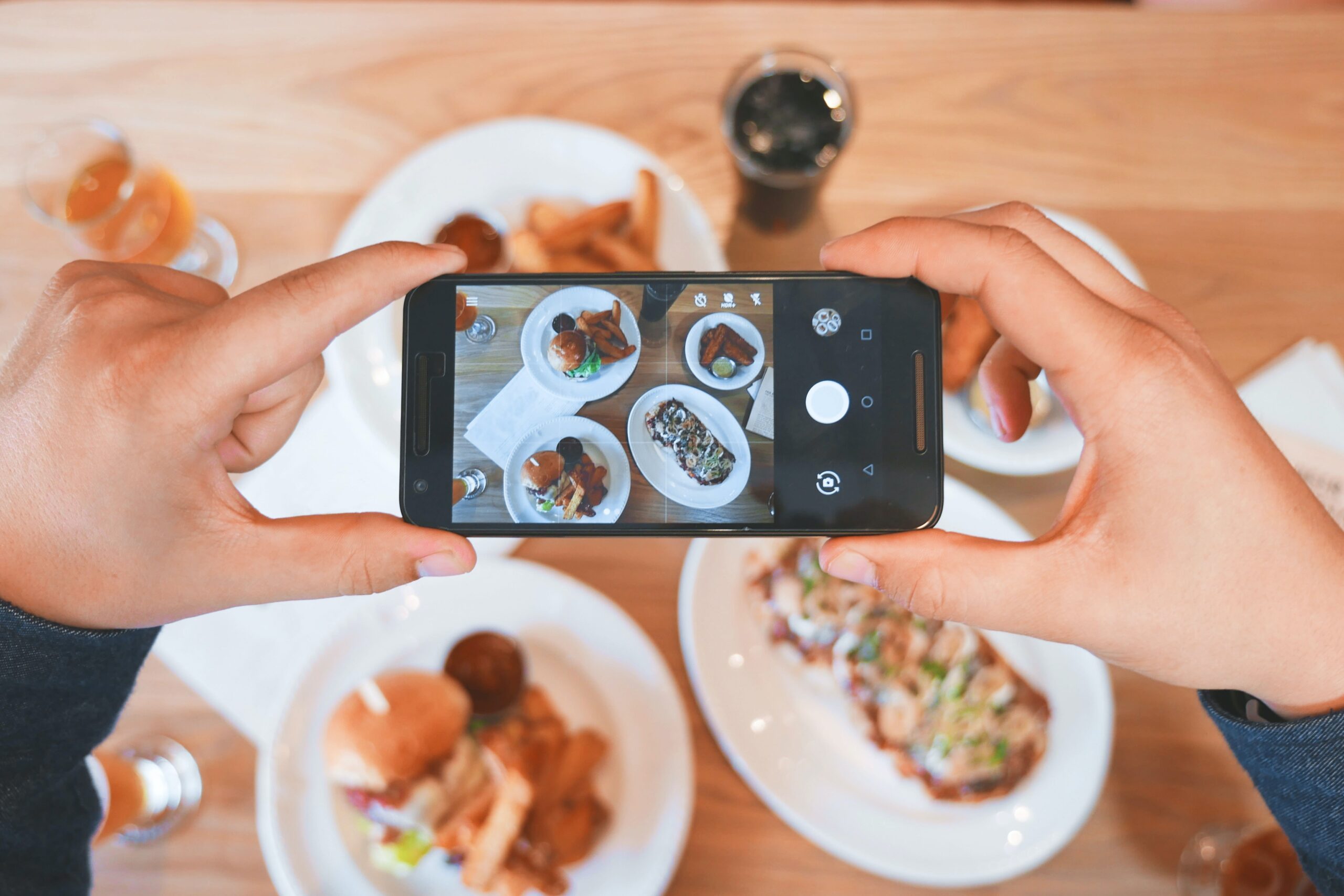 Uma mão segurando um celular apontando a câmera para vários pratos de comida sobre uma mesa de madeira. Foto ilustrativa de marketing digital para restaurantes