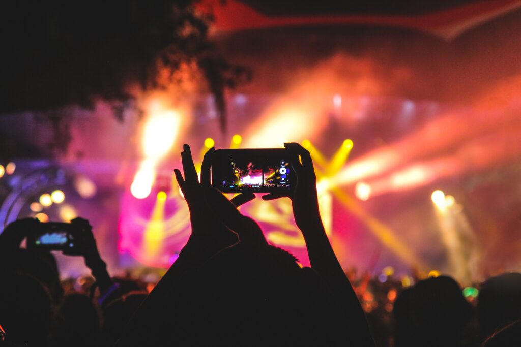 Pessoas seguram celular para tirar foto ou gravar vídeo na plateia de um show, num ambiente bem colorido. Foto ilustrativa da relação da internet com a música, uma das bases do marketing digital musical.