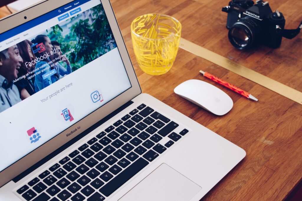 Imagem mostra notebook com tela onde está escrito facebook ads. o computador está em cima de uma mesa de madeira, ao lado de um mouse branco, uma caneta vermelha e um copo de vidro com detalhes amarelos.